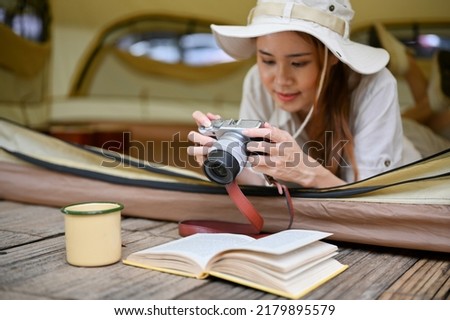 Attractive young Asian female laying in her camping tent, using camera to check her beautiful forest picture. Camping concept