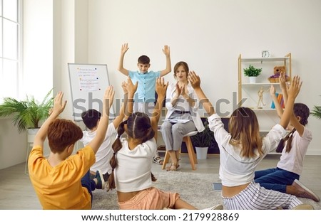 Students and teacher having fun in class. Kids playing charades or Simon Says game at school. Small group of children sitting on floor in classroom, looking at classmate and repeating his movements Royalty-Free Stock Photo #2179928869