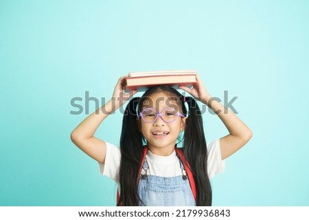 Kid students going to school, girl funny smiling, kid students girl with glasses hold books on her head, blue studio background, school concept.