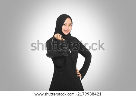 portrait of smiling asian muslim business woman wearing hijab carrying bag while standing isolated on gray background