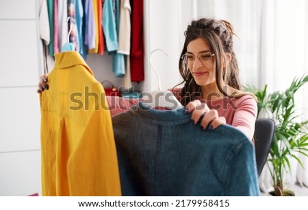 Young happy woman holding two shirts and comparing them, she is choosing what to wear Royalty-Free Stock Photo #2179958415