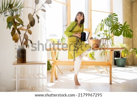 Young woman in apron as housewife or gardener takes care of green plants, watering flower pots in sunny room. Concept of gardening and hobby Royalty-Free Stock Photo #2179967295