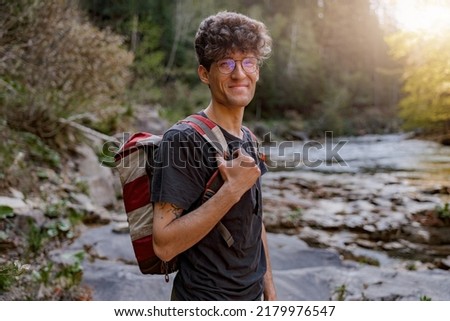 Young cheerful male tourist with backpack standing at river in mountains and smiling.