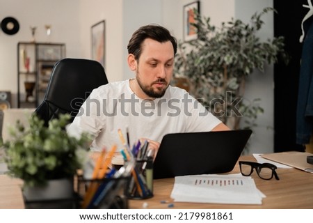 A man is working on his new project on a laptop. He is completely focused on work. There are stationery and sheets of paper on table next to it.