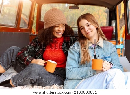 Two happy multiracial girlfriends on car trunk in fall forest, embracing and drinking hot tea during adventure trip in autumn nature, smiling female friends enjoying life while traveling together Royalty-Free Stock Photo #2180000831