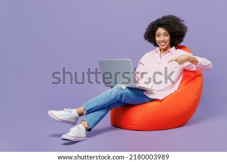 Full body fun young woman of African American ethnicity 20s wear pink striped shirt sit in bag chair hold use work point finger on laptop pc computer isolated on plain pastel light purple background