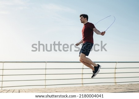 Full size young strong sporty toned fit sportsman man 20s wear sports clothes jumping on skipping rope warm up training at sunrise sun dawn over sea beach outdoor on pier seaside in summer day morning Royalty-Free Stock Photo #2180004041
