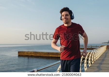 Young smiling strong sporty athletic fit sportsman man wear sports clothes heapdhones listen music warm up training at sunrise sun dawn over sea beach outdoor on pier seaside in summer day morning. Royalty-Free Stock Photo #2180004063