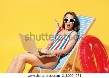 Full body length happy young woman wear red blue one-piece swimsuit sit on chair work hold laptop pc isolated over vivid yellow color wall background studio. Summer hotel pool sea rest sun tan concept