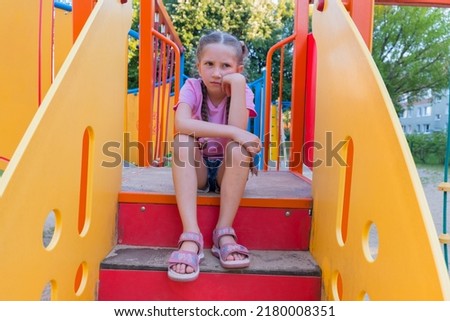 A child girl with blond hair in a pink T-shirt sits offended and sad on the playground.  Royalty-Free Stock Photo #2180008351