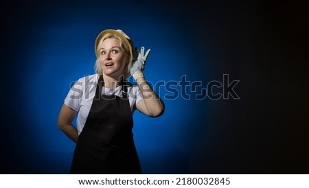 A woman in a white t-shirt, black apron, gloves and a hat put her hand to her ear and eavesdrops on a dark background with copy space.