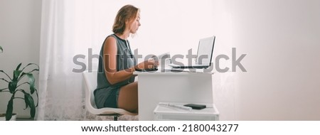 A young woman sitting at a table and leafing through a book. Nearby are a laptop and office supplies. Interior is white color. Wide format. Copy space.