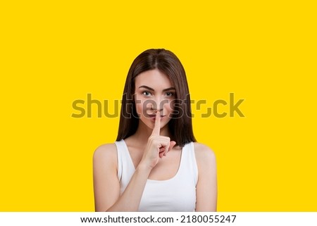 Portrait of an attractive coquettish cute girl holding a finger to her lips in a white top on a yellow background. Secret. Shut up.