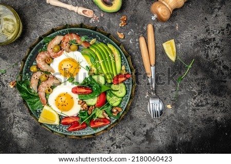 Ketogenic diet breakfast shrimps, prawns, soft fried egg, fresh salad, tomatoes, cucumbers and avocado on a dark background. Keto, paleo lunch. Top view. Royalty-Free Stock Photo #2180060423