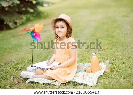 Cute happy little girl in a hat having fun in park meadow. Child sitting on a blanket, drawing picture and and look at camera. Picnic on nature