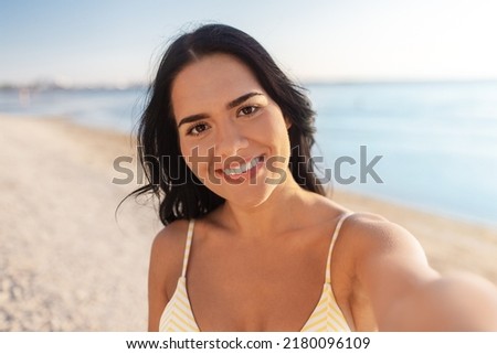 people, summer and swimwear concept - happy smiling young woman in bikini swimsuit taking selfie on beach