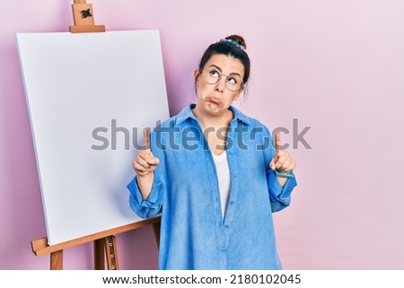 Young hispanic woman standing by painter easel stand pointing up looking sad and upset, indicating direction with fingers, unhappy and depressed. 