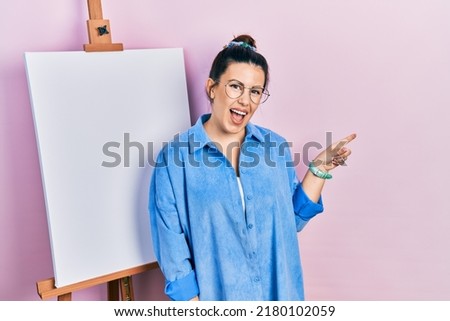 Young hispanic woman standing by painter easel stand with a big smile on face, pointing with hand finger to the side looking at the camera. 