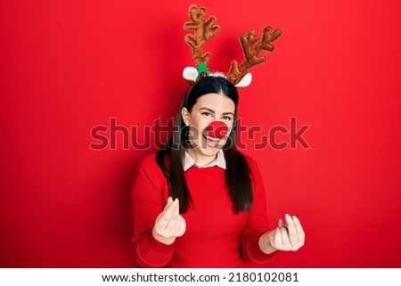 Young hispanic woman wearing deer christmas hat and red nose doing money gesture with hands, asking for salary payment, millionaire business 
