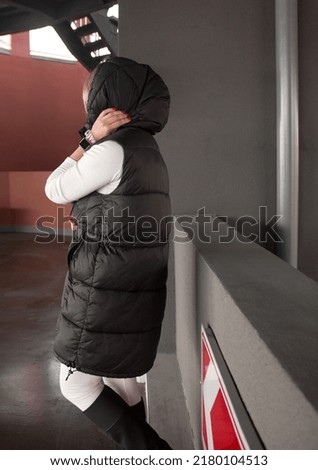 teen walk style. young girl in black long vest, slim white suit and boots is standing casual from the back with hand near hood at parking background and looking apart. lifestyle concept, free space