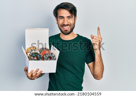 Young hispanic man holding box of tasty colorful doughnuts smiling with an idea or question pointing finger with happy face, number one 