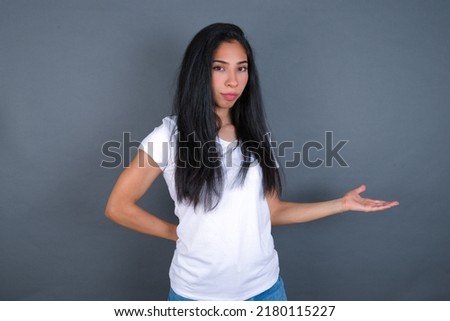 Portrait of young beautiful brunette woman wearing grey T-shirt over white wall  with arm out in a welcoming gesture.