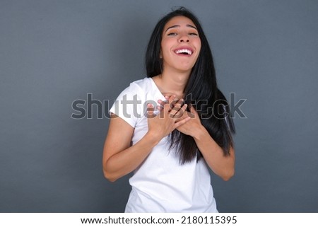 Happy smiling young beautiful brunette woman wearing grey T-shirt over white wall has hands on chest near heart. Human emotions, real feelings and facial expression concept.