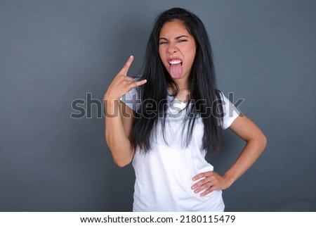 Portrait of a crazy young beautiful brunette woman wearing grey T-shirt over white wall showing tongue horns up gesture, expressing excitement of being on concert of band.