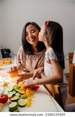 Cute asian little girl kissing her happy smiling mother while they cooking together. Daughter cutting vegetables and mother preparing fresh orange juice