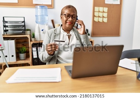 Senior african american woman business worker talking on the smartphone holding credit card at office
