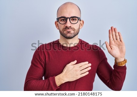 Young bald man with beard standing over white background wearing glasses swearing with hand on chest and open palm, making a loyalty promise oath  Royalty-Free Stock Photo #2180148175