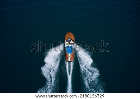 High speed open modern wooden boat moving fast on dark water aerial view. Wooden expensive italian boat with people fast movement on the water top view. Royalty-Free Stock Photo #2180156729