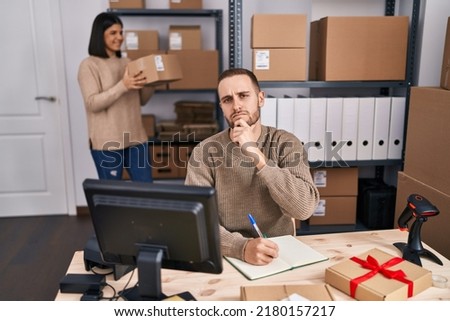 Two young people working at small business ecommerce serious face thinking about question with hand on chin, thoughtful about confusing idea 