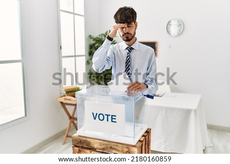 Hispanic man with beard voting putting envelop in ballot box worried and stressed about a problem with hand on forehead, nervous and anxious for crisis 
