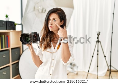 Beautiful caucasian woman working as photographer at photography studio pointing to the eye watching you gesture, suspicious expression 