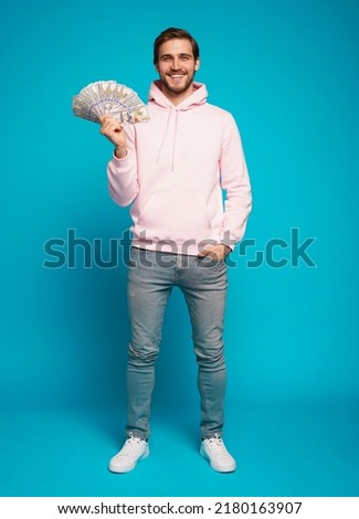 Portrait of a joyful young man holding money cash and celebrating isolated over light blue background.