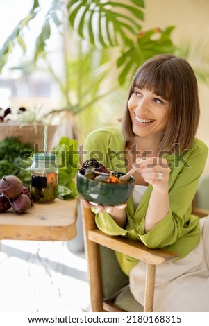 Young cheerful woman eats vegetarian lunch in bowl, sitting by the table full of fresh food ingredients indoors. Healthy lifestyle and wellness concept Royalty-Free Stock Photo #2180168315