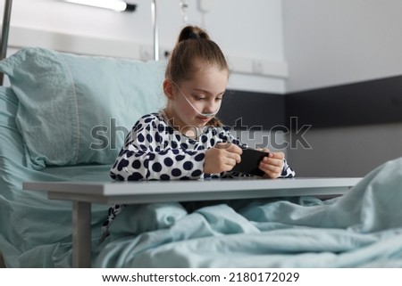 Pretty little sick girl resting on bed while playing games on smartphone. Hospitalized ill kid watching cartoons on phone while sitting inside pediatric healthcare clinic ward.