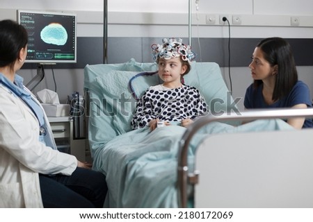Hospitalized sick girl wearing EEG headset resting in patient bed while doctor analyzing brain condition. Neurology specialist examining neurological disease state of ill kid. Royalty-Free Stock Photo #2180172069