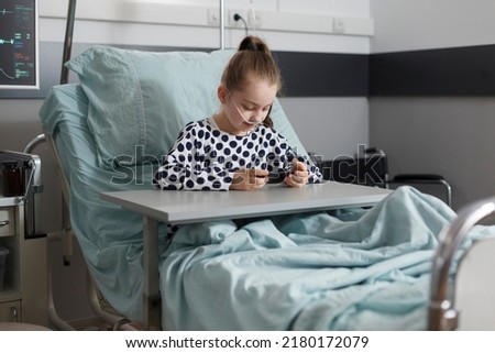Sick girl under treatment resting on patient bed while watching cartoons on smartphone. Hospitalized ill kid playing games on modern mobile phone while resting inside hospital pediatrics ward. Royalty-Free Stock Photo #2180172079