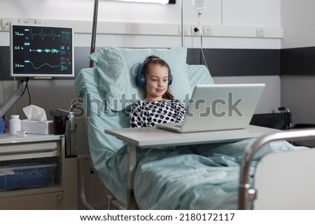 Young sick patient under treatment watching cartoons on laptop while resting on patient bed inside healthcare facility pediatrics ward room. Ill little girl with computer enjoying internet content