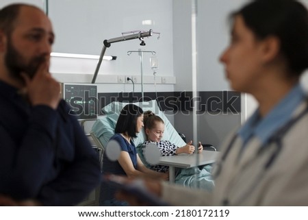 Sick girl playing games on smartphone with mother while father talking with expert pediatrist. Hospitalized ill little kid watching cartoons on mobile phone while resting in hospital pediatric ward Royalty-Free Stock Photo #2180172119
