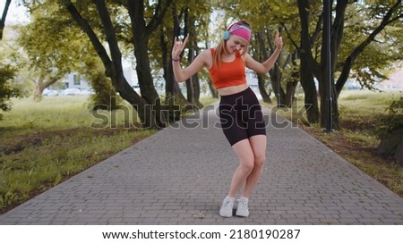 Athletic fitness sport runner girl training exercise, listening music on headphones dancing to camera having fun. Workout cardio in park. Young woman jogger enjoying funny dance. Active sportswoman Royalty-Free Stock Photo #2180190287