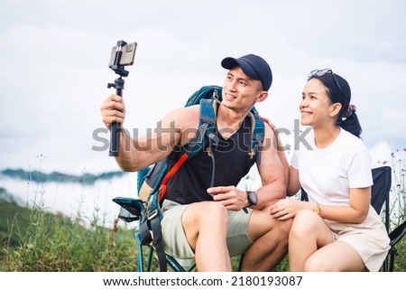 Asian tourist couple with backpack taking selfie photo on vacation trip. Happy traveler man and woman shooting video with selfie stick on mountain and fog background. Blogger and vlog concept.