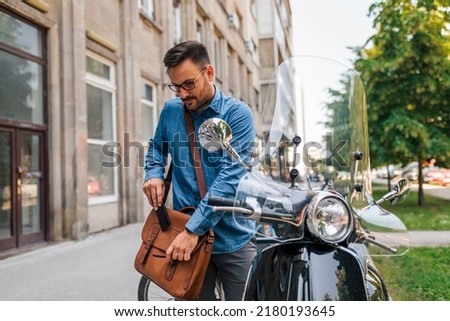 Smiling businessman closing bag while preparing to drive his motor scooter. Male professional standing by parked vehicle on sidewalk. He is commuting in the city. Royalty-Free Stock Photo #2180193645