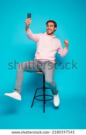 Online Profit. Portrait of a joyful young man sitting on chair and holding mobile phone isolated over light blue background, celebrating financial success Royalty-Free Stock Photo #2180197541