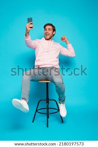 Online Profit. Portrait of a joyful young man sitting on chair and holding mobile phone isolated over light blue background, celebrating financial success Royalty-Free Stock Photo #2180197573