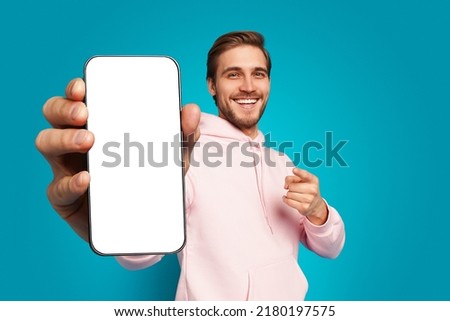 Mobile App Advertisement. Handsome Excited Man Showing Pointing At Empty Smartphone Screen Posing Over Light Blue Studio Background, Smiling To Camera. Check This Out, Cellphone Display Mock Up