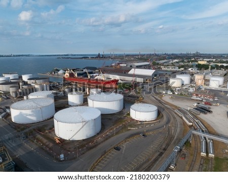 An aerial view of a white petroleum or oil tank field located next to a port. Pictured during the day while a cargo vessel is stationary.