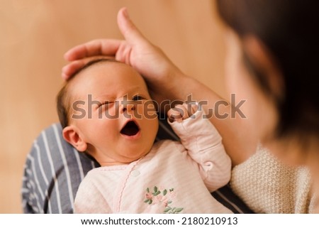 Cute funny infant baby girl making faces lying on mother hands in room at home and try to fall asleep. Motherhood. Woman holding newborn  Royalty-Free Stock Photo #2180210913
